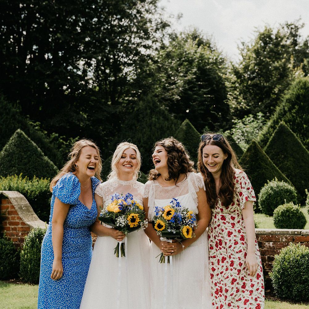 Brides laugh together with two wedding guests in flower and polka dot dresses as they hold their sunflower and gypsophila bouquets