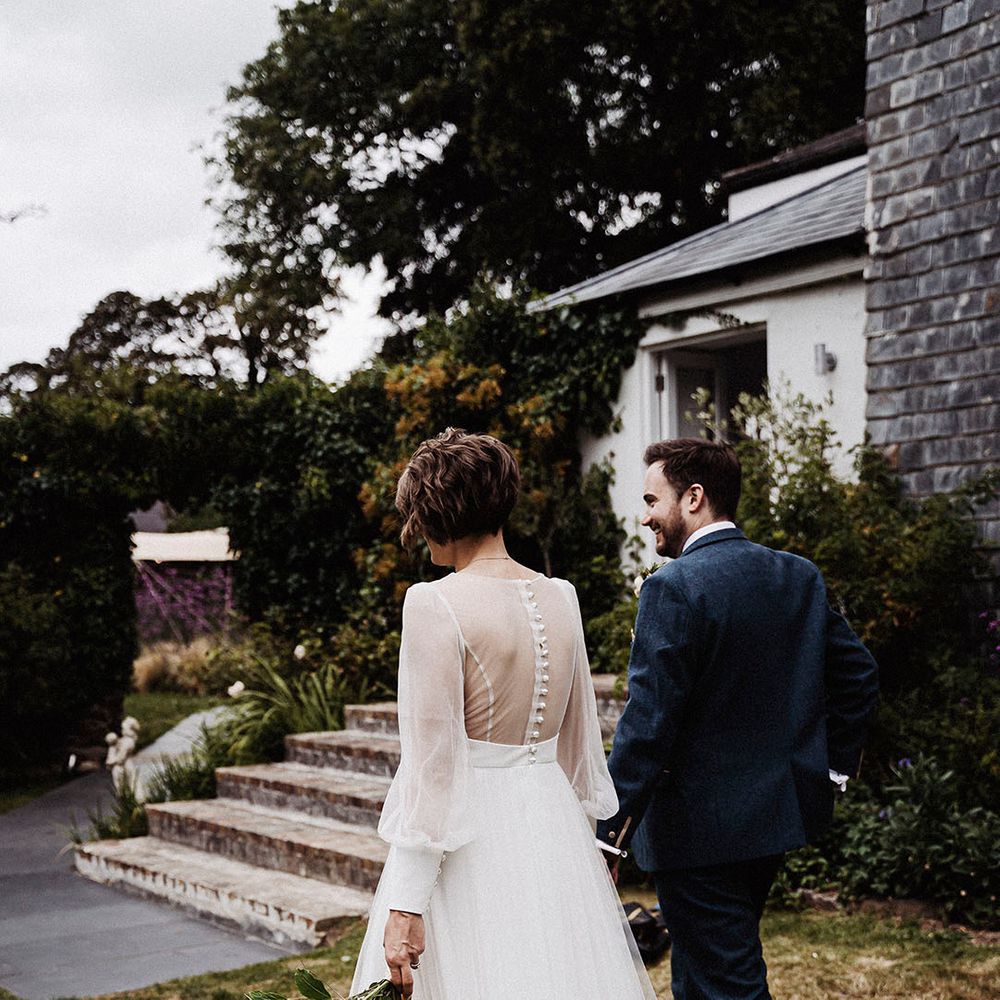 Groom in blue suit walks holding hands with bride in sheer long sleeve wedding dress 