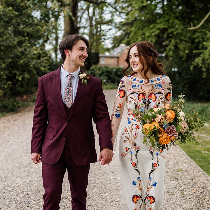 Groom in three piece burgundy suit with bride in colourful flower wedding dress with colourful bouquet 