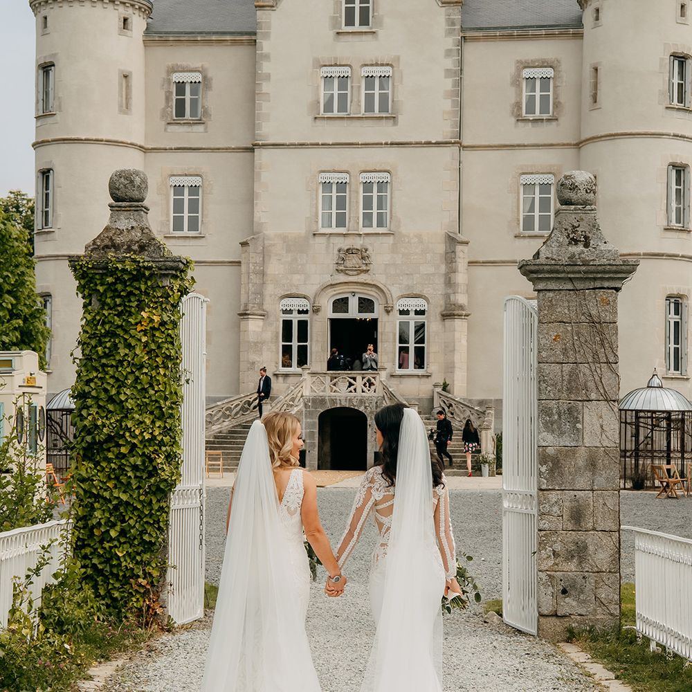 Brides walk through white iron gate together holding hands in front of the chateau