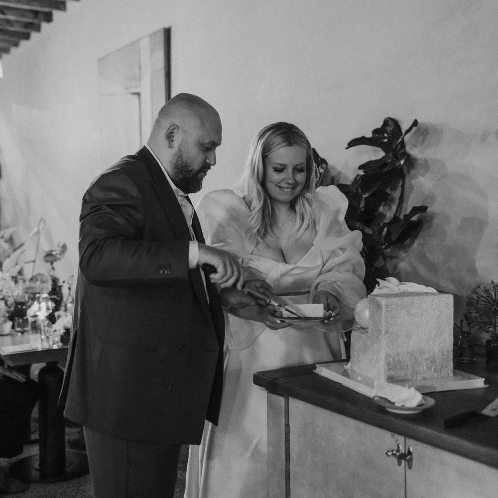black and white portrait of the bride in a wedding dress with long puffy sleeves cutting the square wedding cake 