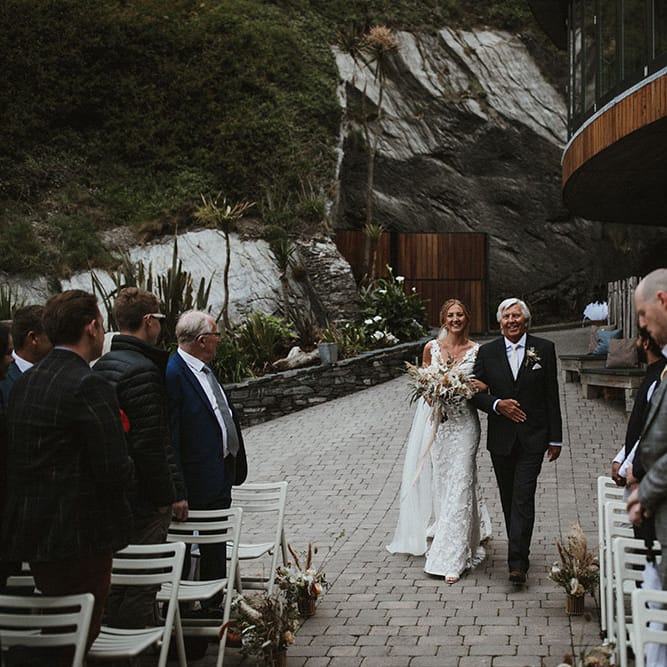 Bride wears lace Enzoani wedding dress as she walks down the aisle with her father who wears black suit and floral buttonhole 