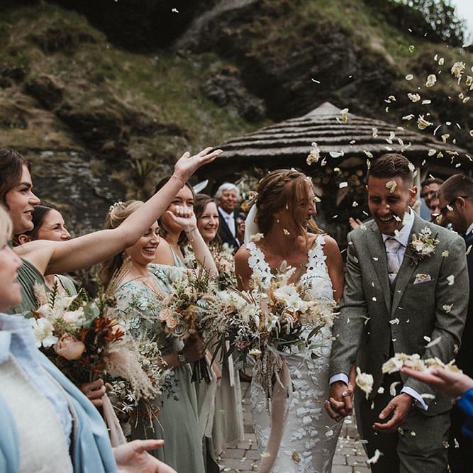 Bride wears lace embellished wedding dress whilst walk through confetti petals with her groom after outdoor wedding ceremony