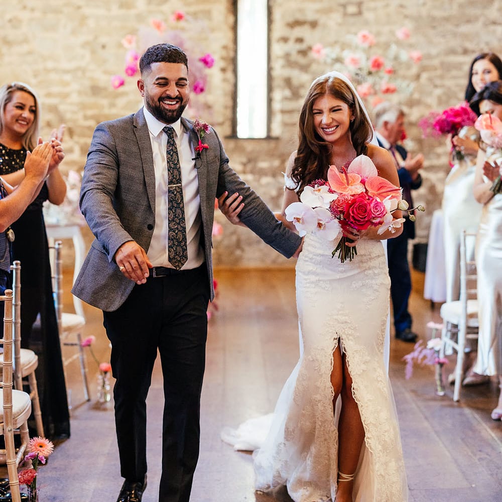 Bride carrying bright pink floral bouquet walks down the aisle with her groom after wedding ceremony at the Priston Mill