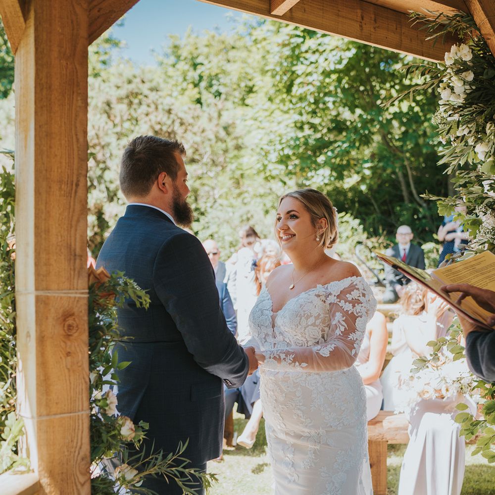 Bride in long sleeve lace wedding dress looks lovingly at her groom outdoors during ceremony at Watermouth Cove