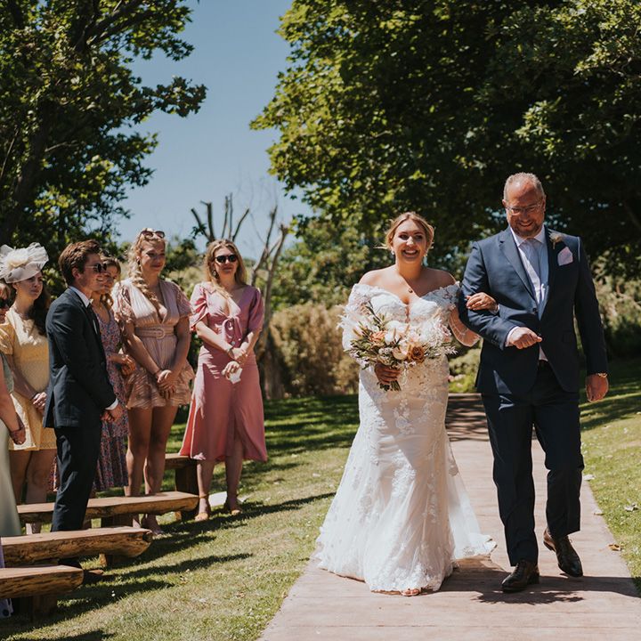 Bride in long sleeve lace wedding dress holding floral bouquet walks down the aisle outdoors at Watermouth Cove