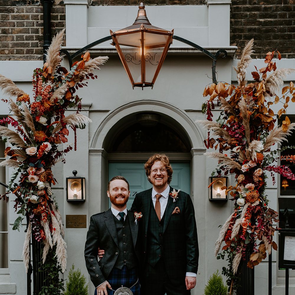 Groom in dark suit with orange tie and autumnal boutonniere standing with best man wearing dark suit, waistcoat, autumnal boutonniere and dark blue tartan kilt underneath large floral arch with roses, peonies, foliage and pampas grass
