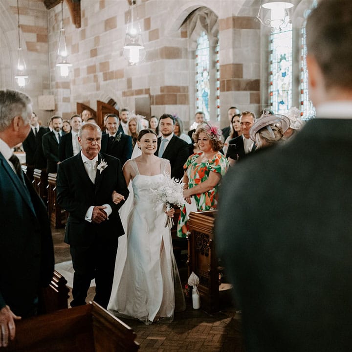 Father of the bride in dark suit with paisley silver tie walks the bride down the aisle for church ceremony 