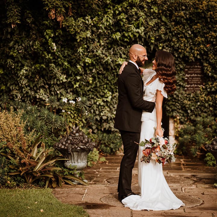 Groom in classic black tuxedo with black bowtie embracing bride in v neck satin wedding dress with sheer butterfly sleeves and ruffled layer on top holding wedding bouquet with white and pink carnations, garden roses, red strawflower, eucalyptus, baby’s breath and various dried flowers
