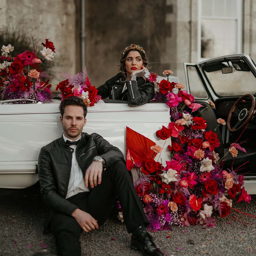 Bride sitting in off white classic convertible wedding car with red roses, off white peonies, lilac dried lowers, dahlias, orchids, roses and amaryllis wearing embroidered black leather jacket  and groom sitting outside it wearing white shirt, black bowtie, black suit trousers and slim fitting black leather jacket 