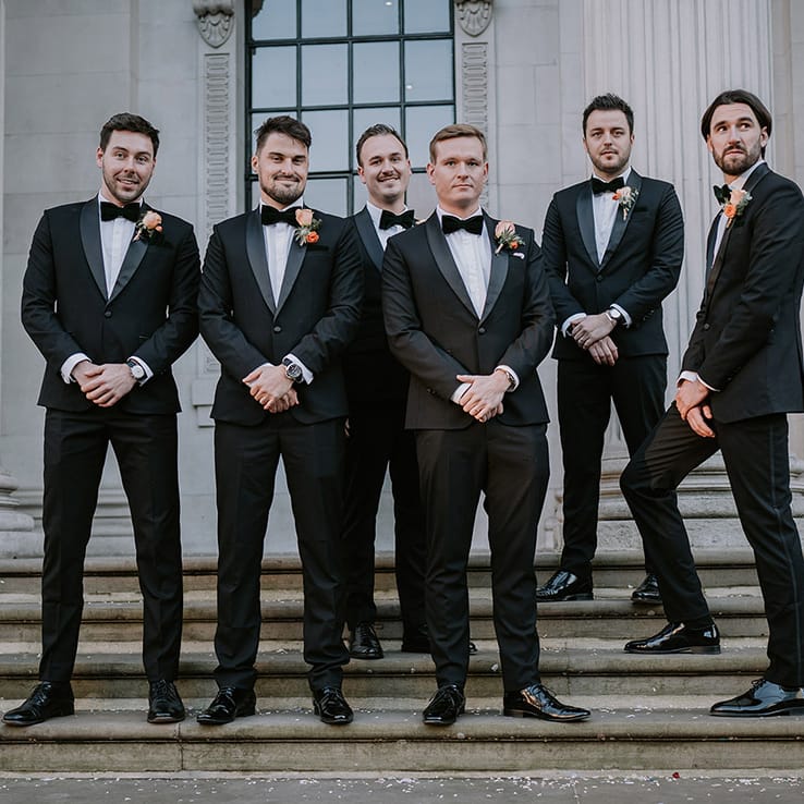 Groom in stylish black tuxedo with black bowtie and light orange rose boutonniere standing with groomsmen in matching black tuxedos 