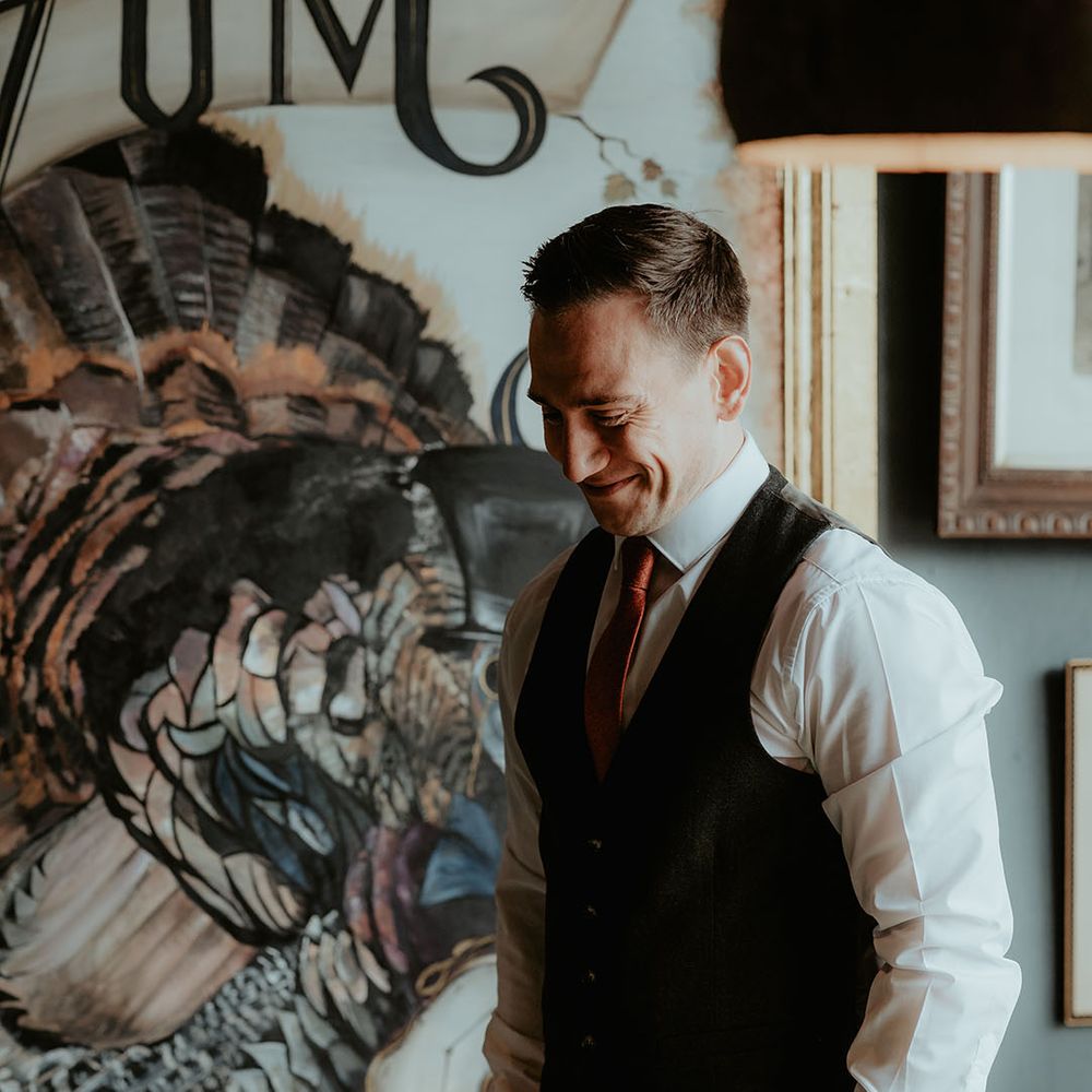 The groom smiles in a black trouser and waistcoat with a white shirt and an orange tie 