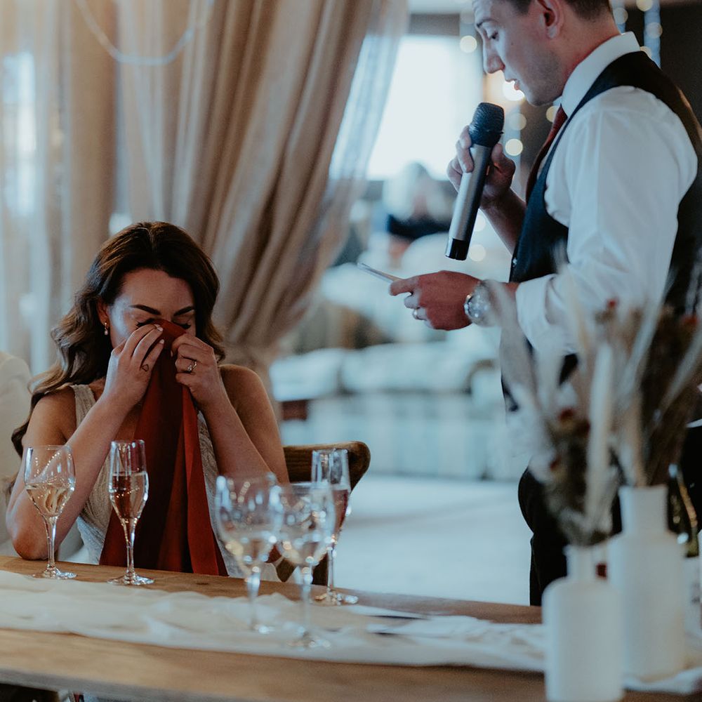 The groom stands up holding a microphone and papers for his speech with the bride sitting down wiping her eyes with the napkin as she cries 
