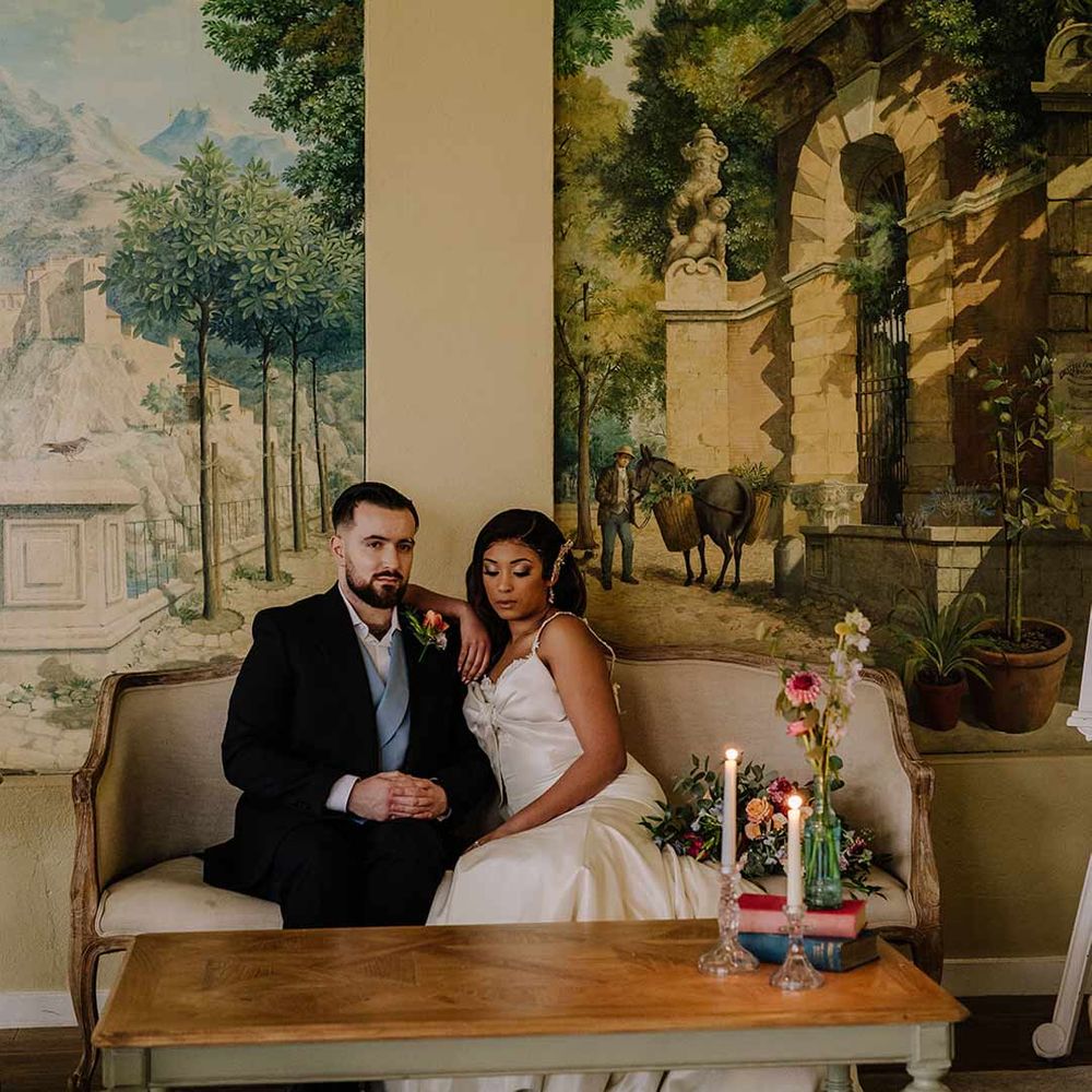 Bride in bow front sleeveless satin wedding dress with large gold bridal hair clip sitting with groom in black blazer with pink garden rose and foliage boutonniere, cornflower blue waistcoat on chaise lounge at Braxted Park wedding venue 