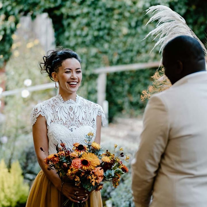 Bride in white mesh lace bridal crop top and yellow wedding skirt holding dried autumnal wedding bouquet with sunflowers, garden roses, pampas grass, eucalyptus, yellow tall kangaroo paws, orange osmanthus flowers, foliage and dried flowers and smiling at groom at Broadfield Court
