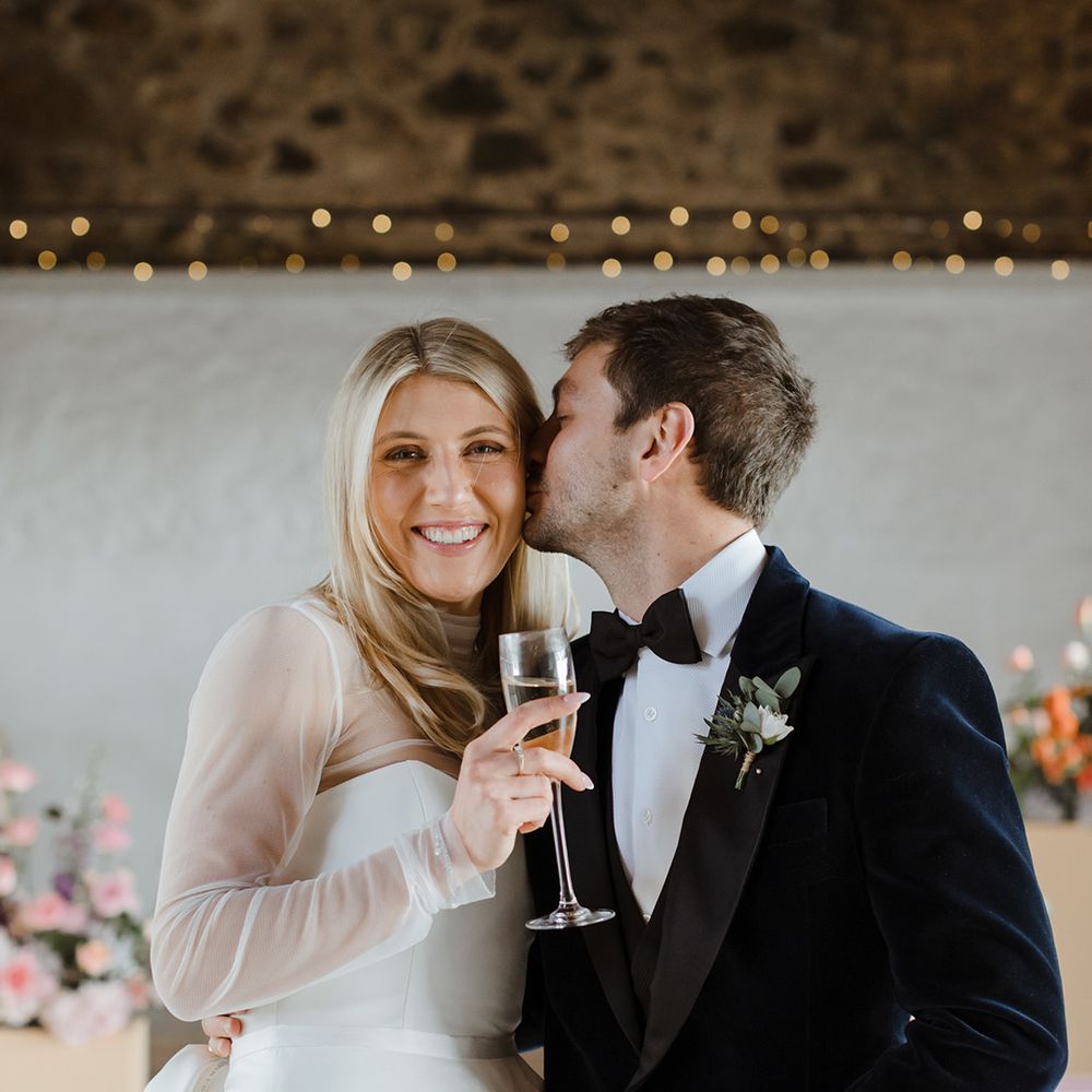 Groom in black tie kisses the bride on the cheek who holds a champagne glass