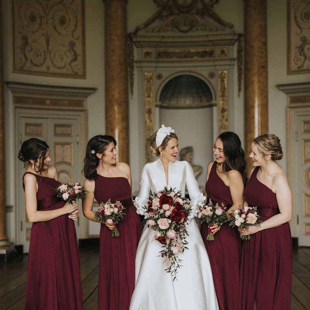 Bride in classical long sleeve satin wedding dress standing with bridesmaid in one shoulder burgundy bridesmaid dresses at Stowe House wedding