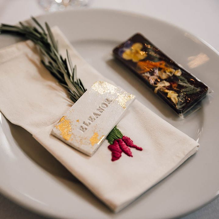 Wedding place setting with white plated decorated with embroidered wedding napkin, sprig of rosemary, and gold flake wedding place name 