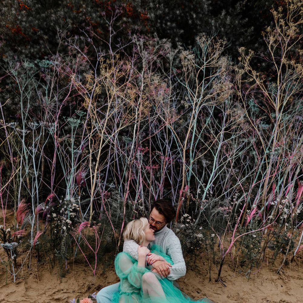Bride in a long sheer green wedding dress barefoot on the beach embracing her husband 