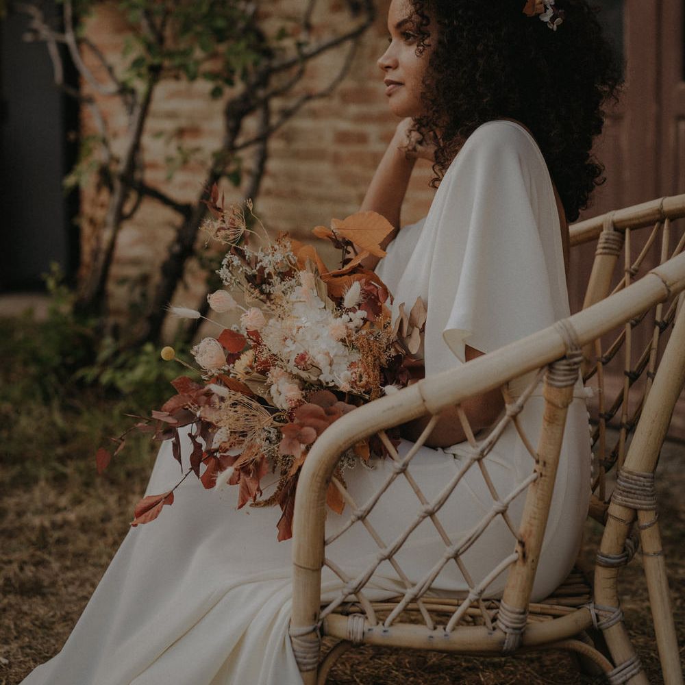 Beautiful bride in a crepe wedding dress with naturally curly hair sitting on a wicker chair holding an autumn leave wedding bouquet 
