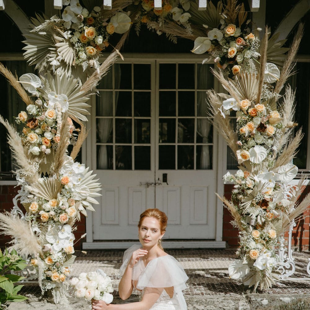 Bride sitting under boho inspired flower arch wearing a floral lace wedding dress with tulle shoulder detail