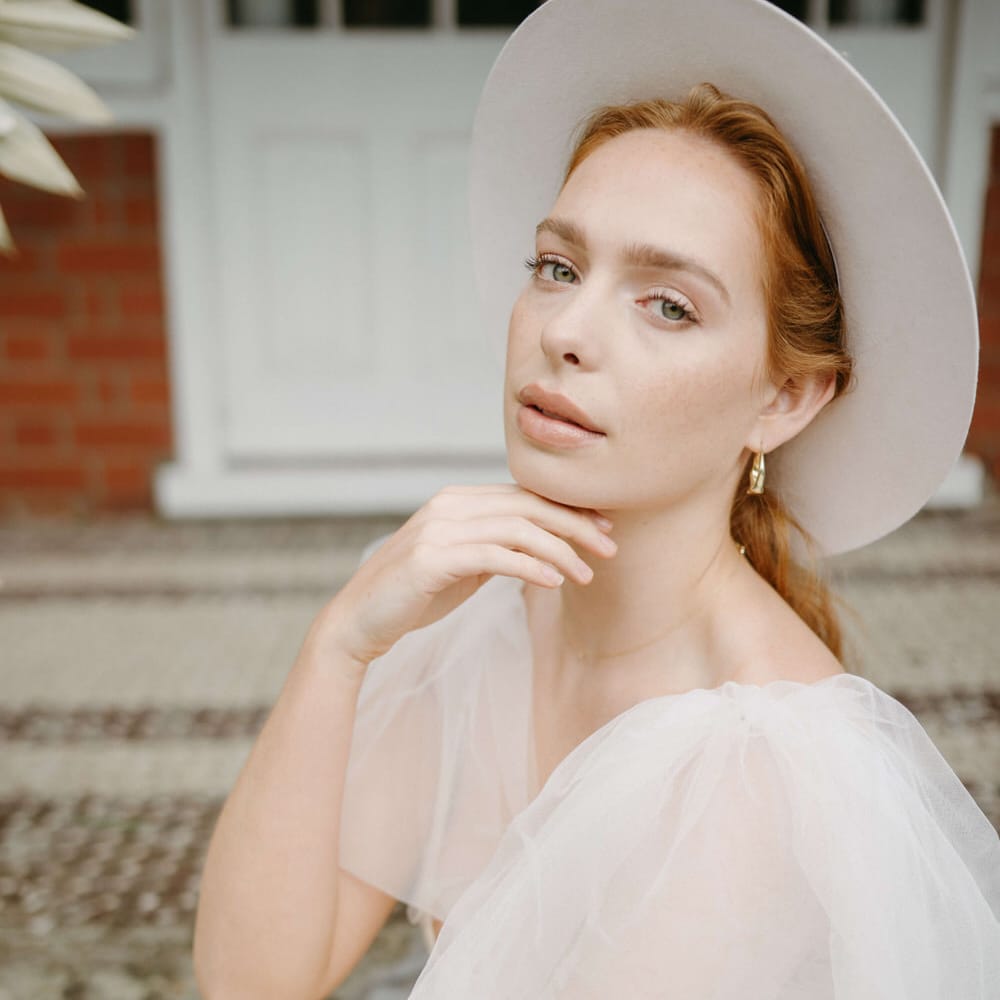 Bride wearing a blush pink stetson hat, puffed tulle shoulder wedding dress with plunge neckline and natural makeup, holding a bouquet of peonies