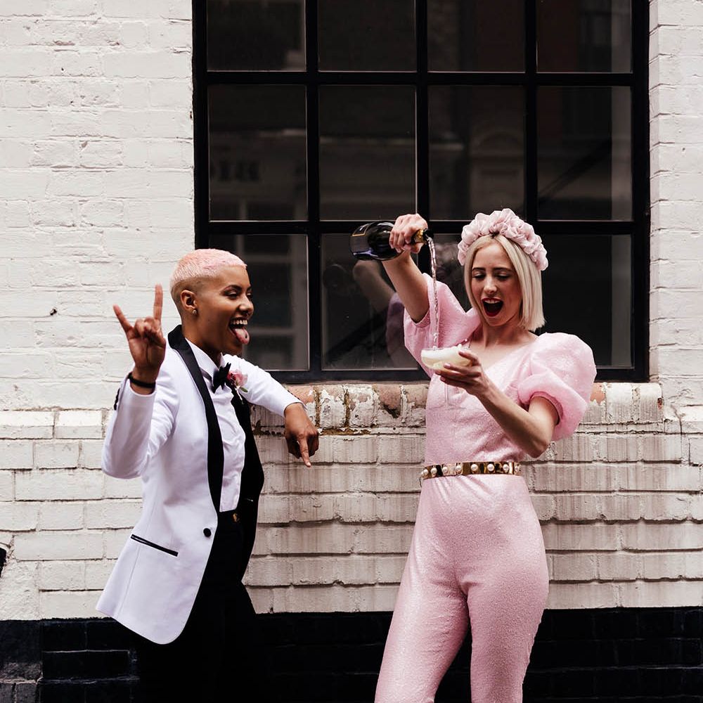 Bride in a shimmering pink jumpsuit with peg leg trousers and puff sleeves during champagne for her bride in a white tuxedo 