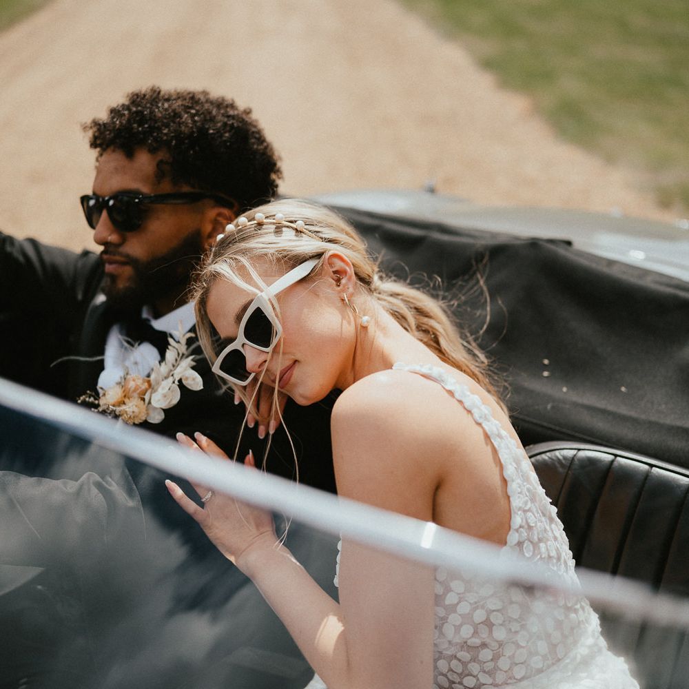 Bride in an appliqué wedding dress, peal headband and white sunglasses resting her head on her grooms shoulder as they sit in their vintage wedding car 