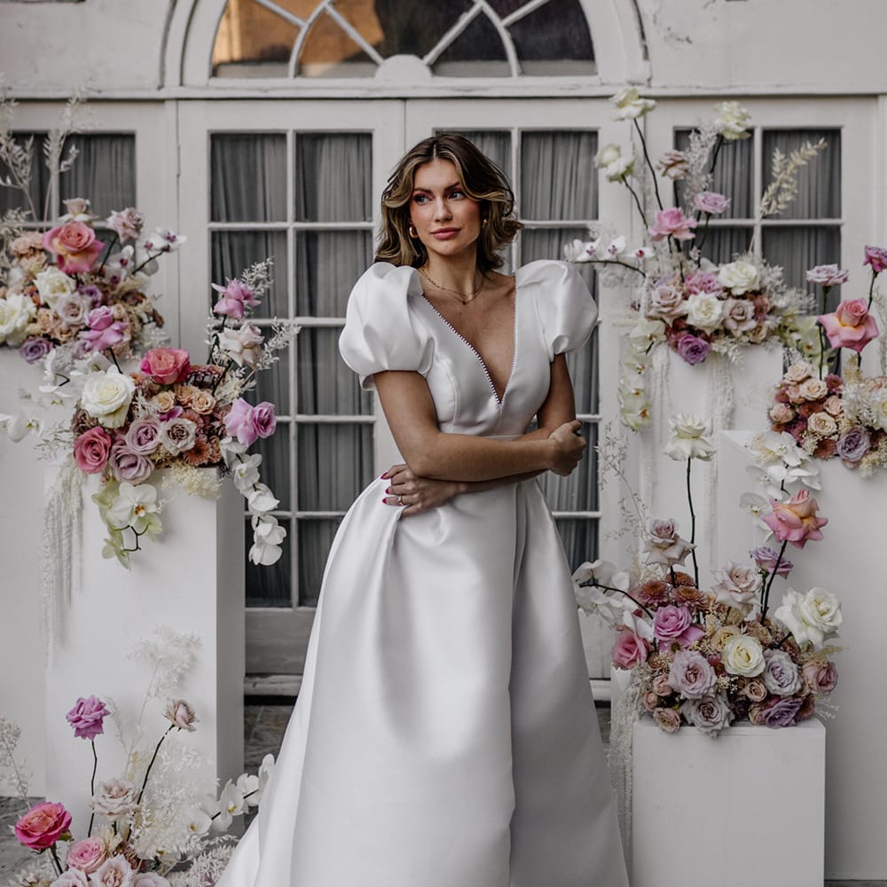 Stylish bride in a plunging neckline wedding dress with puff sleeves standing in front of the wedding ceremony floral arrangements with pink and white orchids and roses 