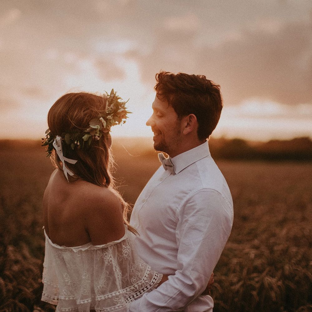Bride in Grace Loves Lace bardot wedding dress and flower crown stands with groom in white shirt by wheat field at golden hour at Isle of Wight wedding with macrame wedding decor