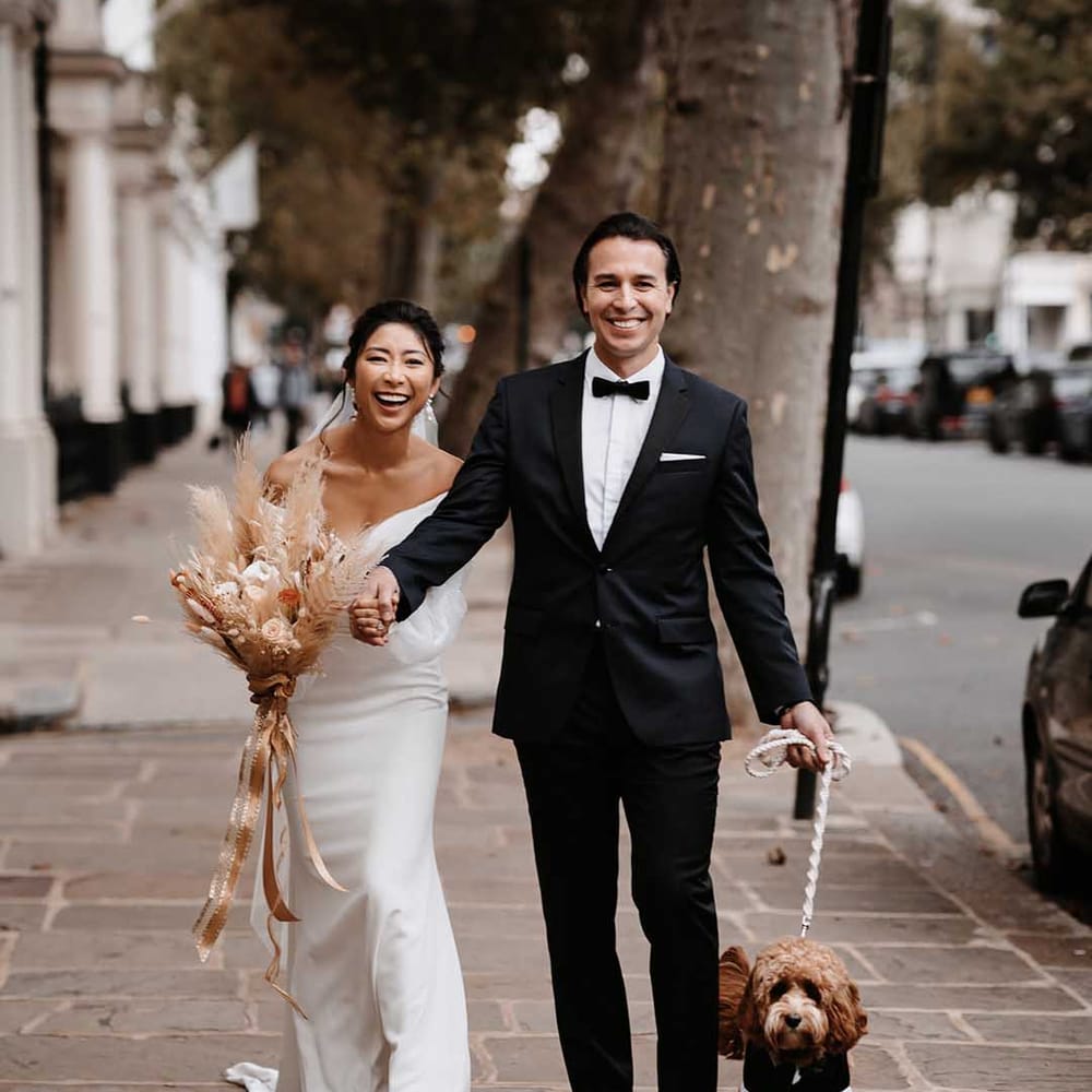 Bride in simplistic wedding dress with nude toned heels holding hands with Groom in sleek black tux and cockapoo in a tux 
