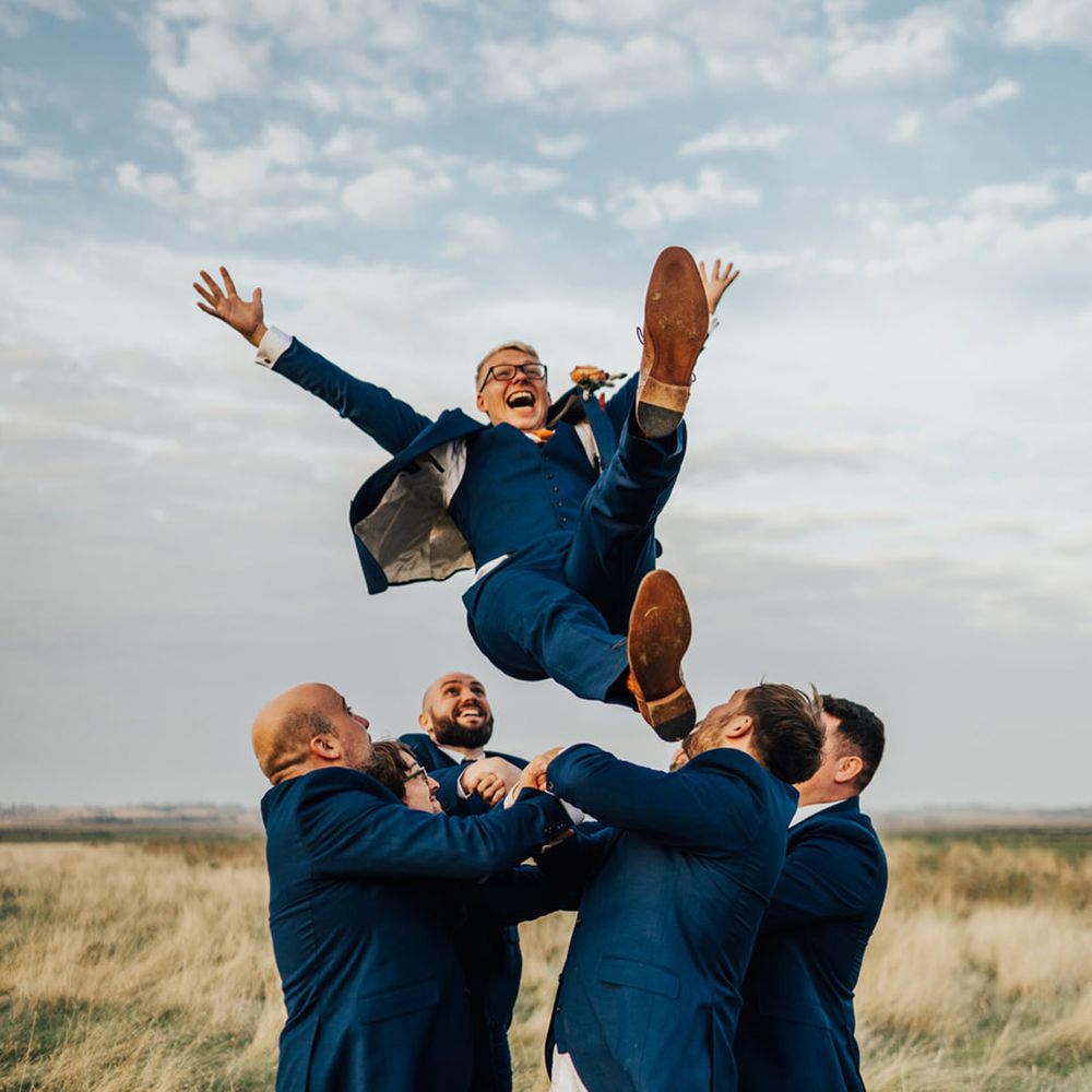 Groomsmen in Navy suits & tanned belts throw Groom in the air in open field & laugh