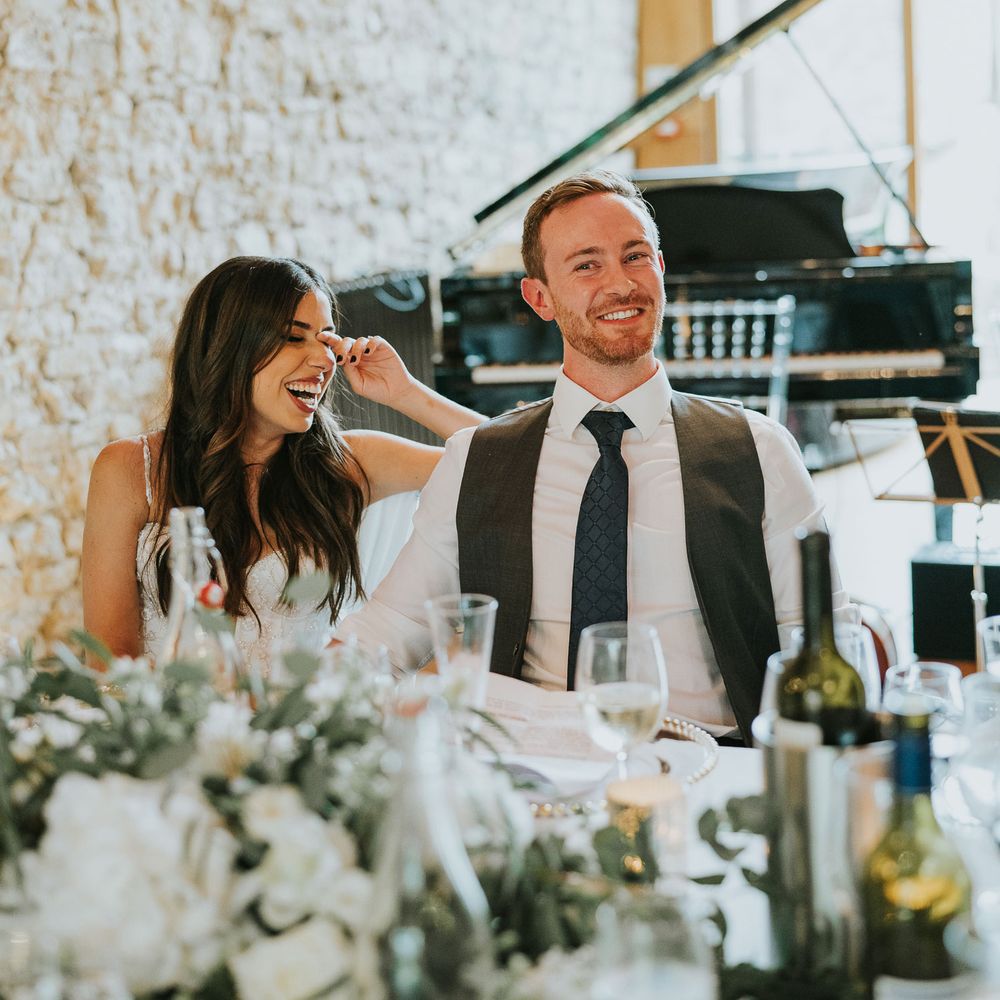Laughing bride in Pronovias wedding dress sits holding hands with groom in white shirt and grey waistcoat during speeches at wedding reception