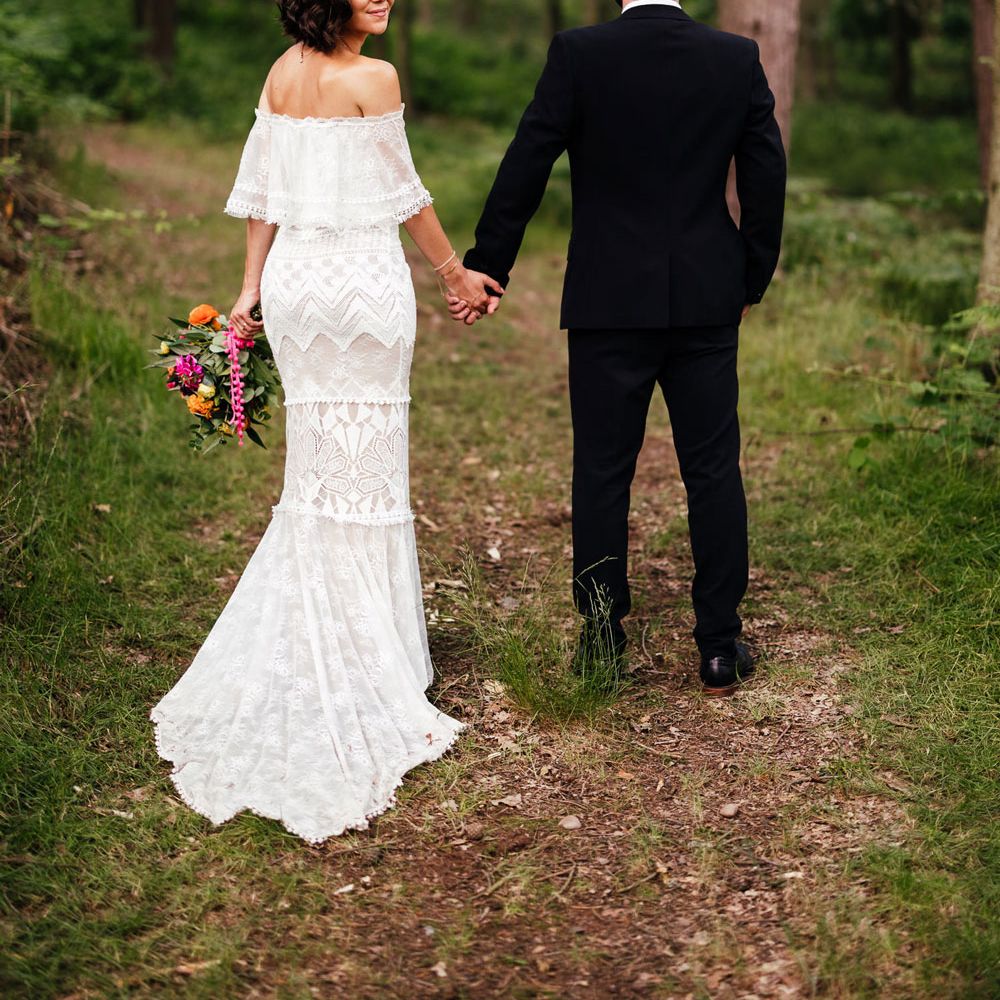 Bride in off the shoulder Grace Loves Lace wedding dress and flower crown holds colourful wedding bouquet as she looks over her shoulder whilst walking through woodland with groom in black suit
