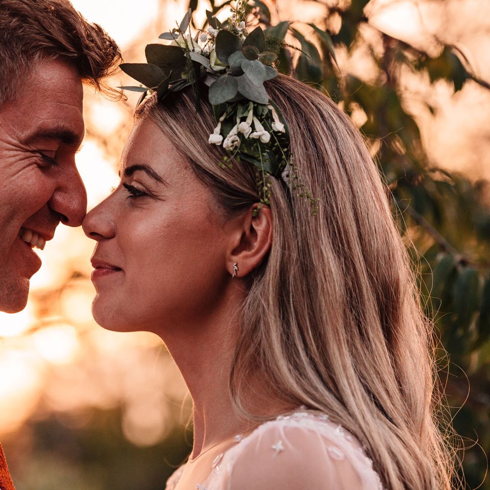 Bride in mesh sleeve wedding dress and boho bridal flower crown stands nose to nose with groom in white shirt and red tie during golden hour