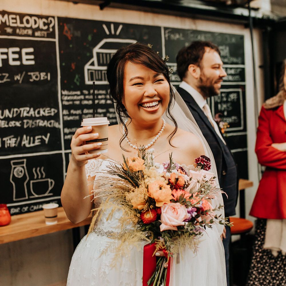 Bride in lace off the shoulder wedding dress and pearl necklace holding bright mixed orange and pink bouquet laughs whilst holding disposable coffee cup 