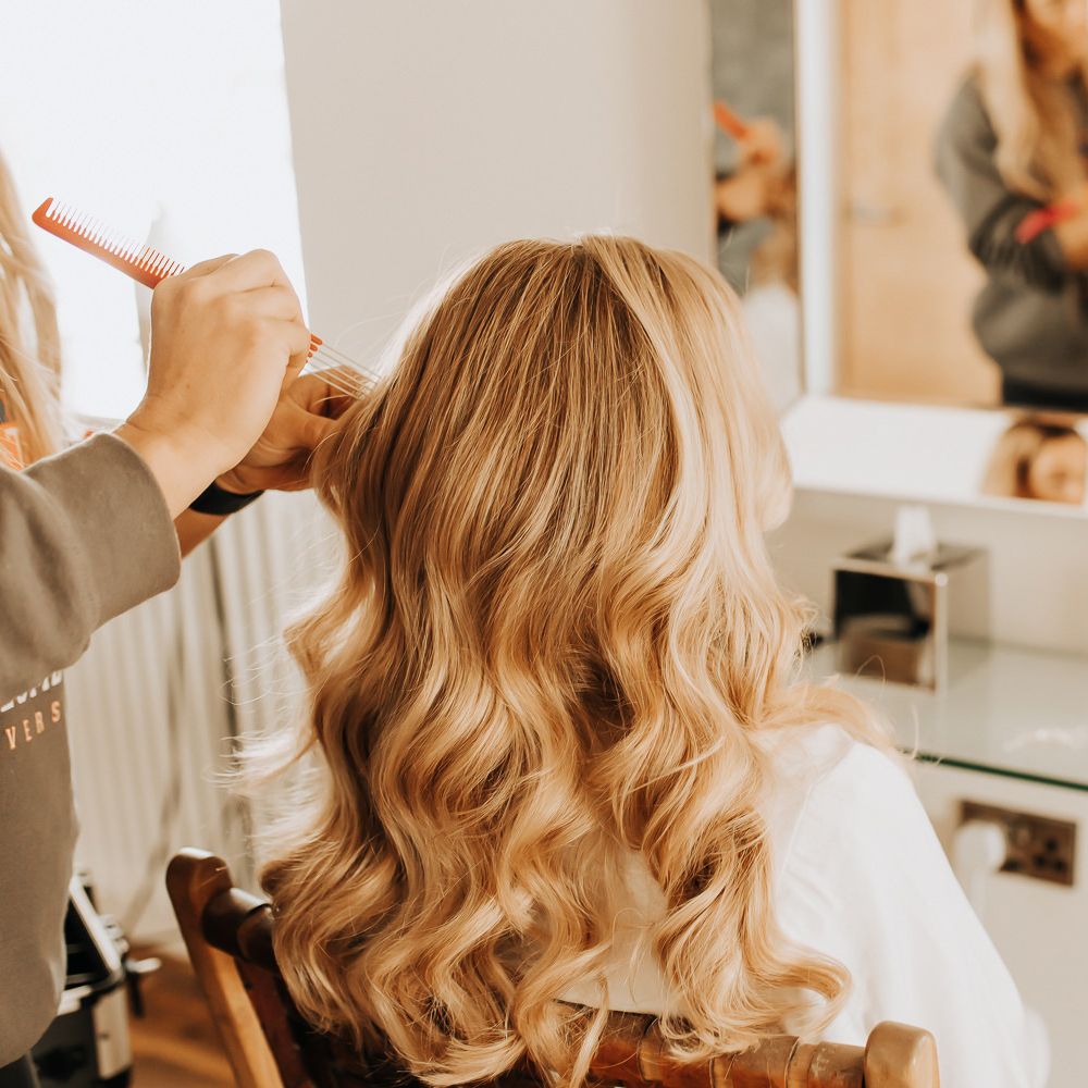 Bride with blonde curled hair sits having her hair styled before wedding
