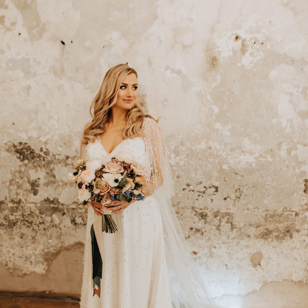 Bride in white wedding dress with pearl mesh overlay stands holding mixed pastel wedding bouquet 