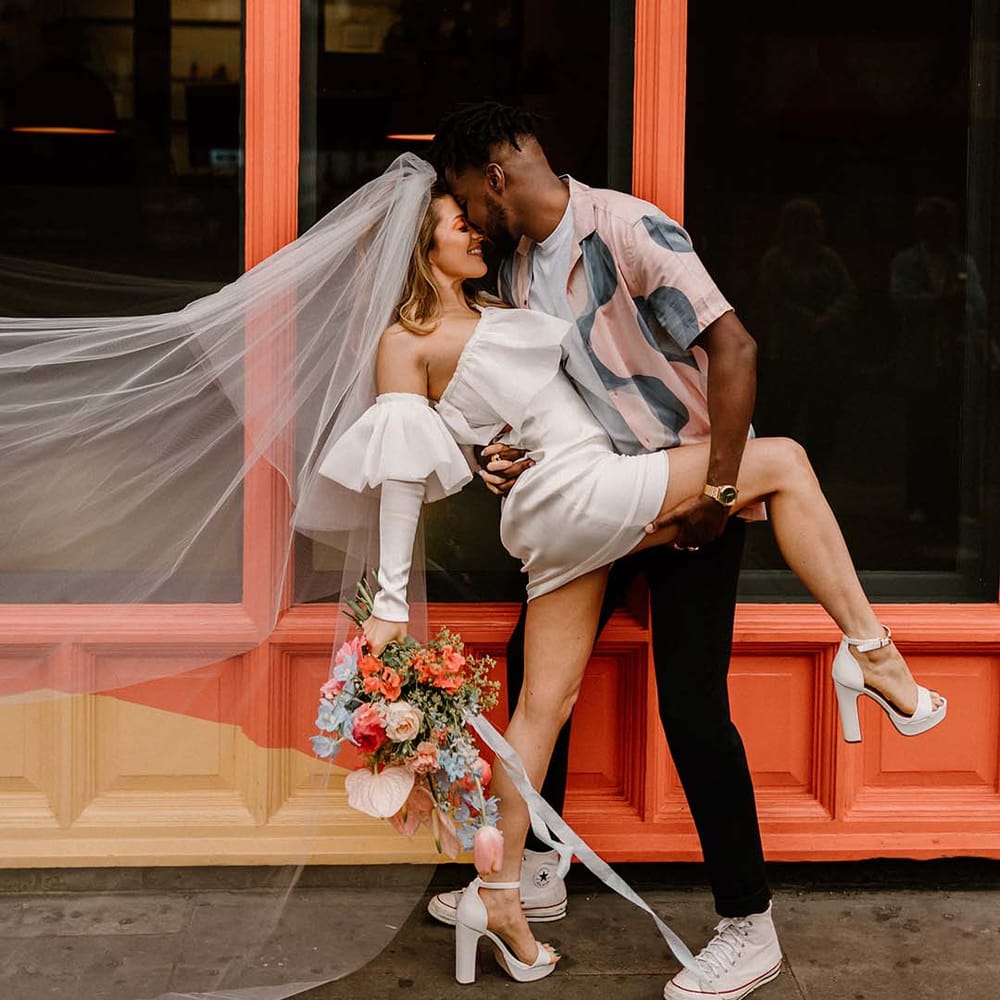 Groom in a patterned shirt leaning his bride over for a kiss in a short wedding dress and long veil at their stylish city elopement 