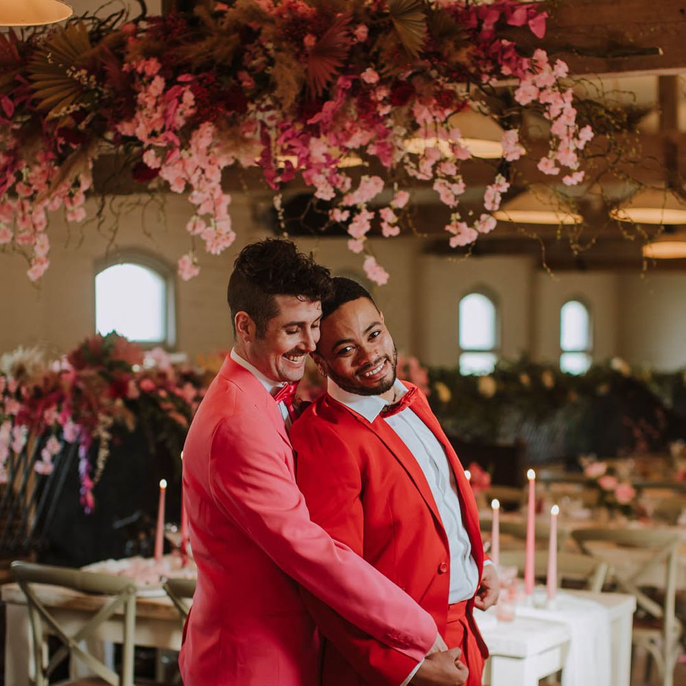 Groom in a pink suit and white boots embracing his partner in a red suit under a wooden beam decorated with cherry blossom and pink flowers 