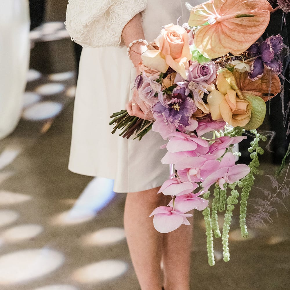 Bride holding orange and purple wedding bouquet with orchids and roses wearing short wedding dress and velvet platform block heels