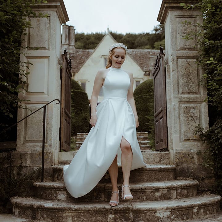 Bride in halter neck wedding dress with pleated skirts, a pearl crown and pearl embellished wedding shoes walks down some stairs 