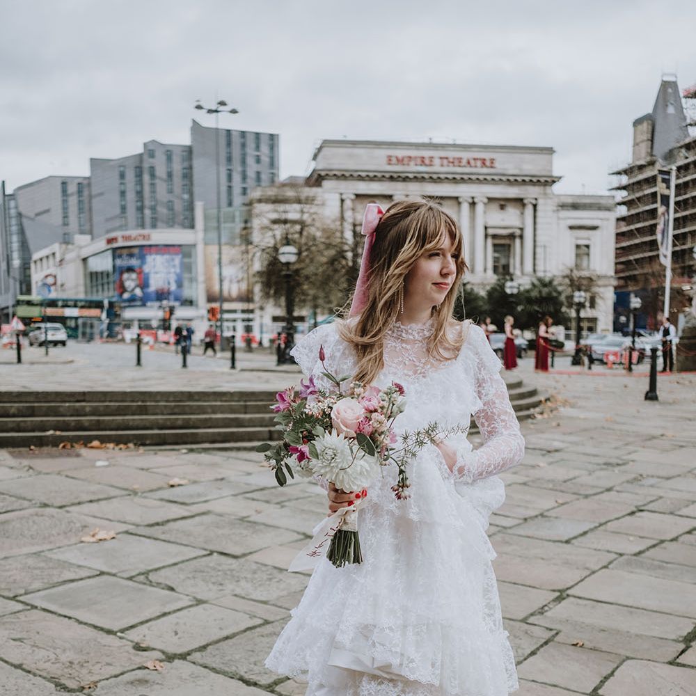 Bride wears ruffled lace vintage wedding dress from Oxfam with long sleeves and pink bow in her long blonde hair 