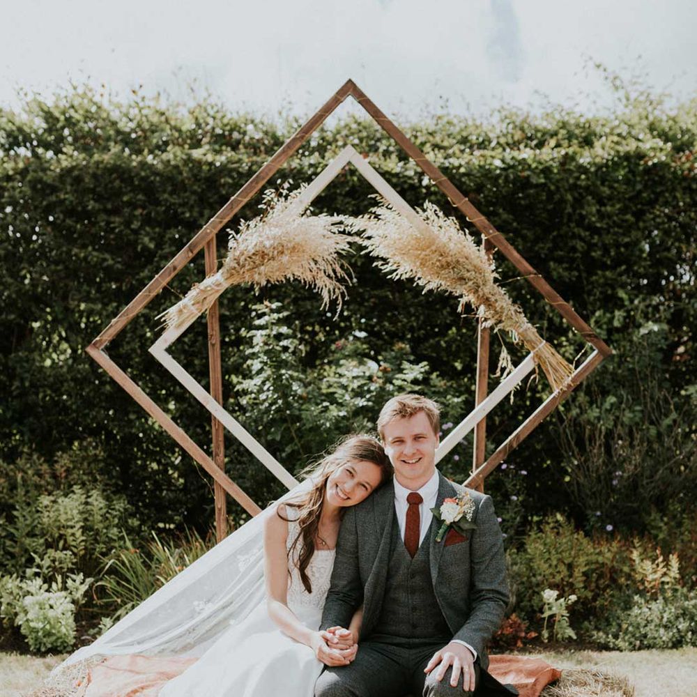Bride in Bardot top wedding dress and veil and groom in dark suit sitting by DIY geometric wedding decoration