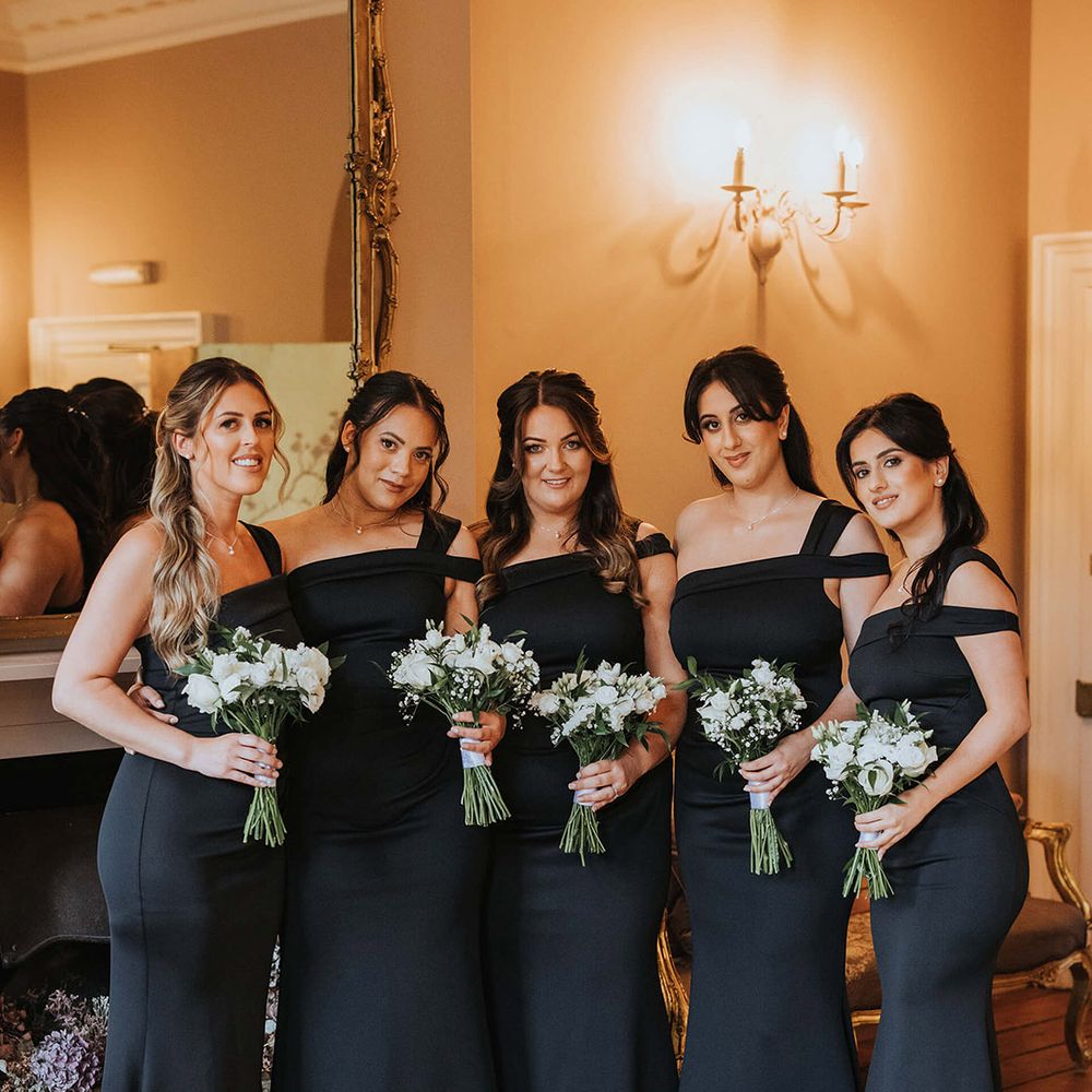 Bridesmaids in asymmetric one shoulder black bridesmaid dresses for the black tie wedding holding white flower bouquets 