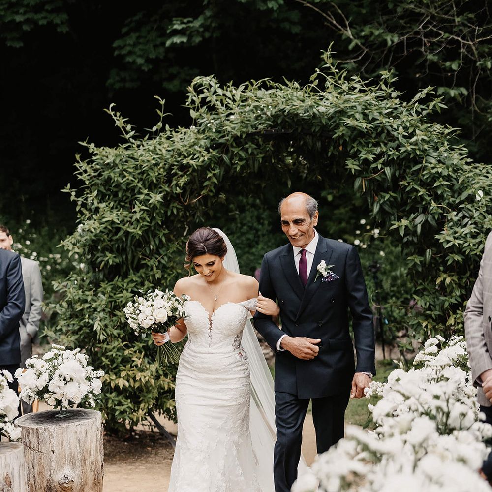 Father of the bride in black double breasted jacket with dark purple tie walking the bride down the aisle in a floral lace wedding dress 