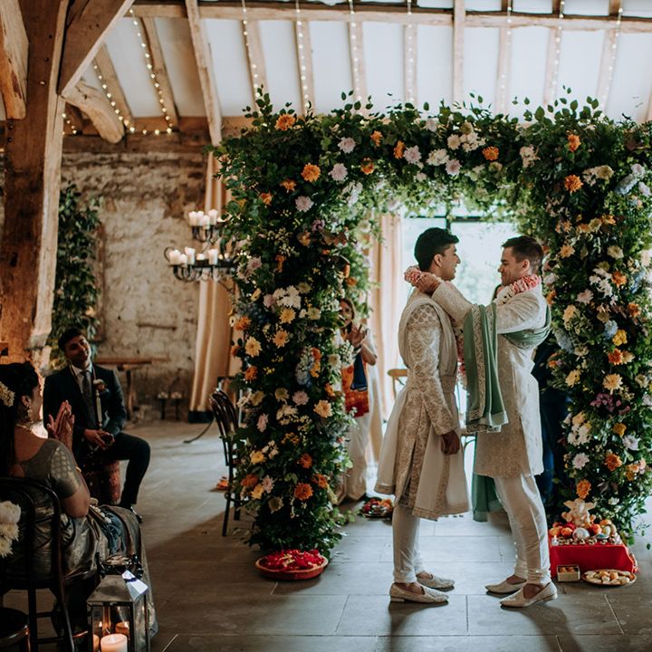 Groom places lei around his grooms neck as they stand beneath floral arch during barn wedding 