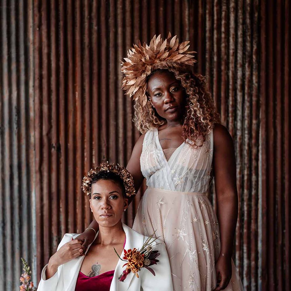 Bride in white silk suit with autumnal dried floral boutonniere sitting on red velvet seat with bride in sleeveless floaty peach gown with embroidered stairs and gold leaf crown