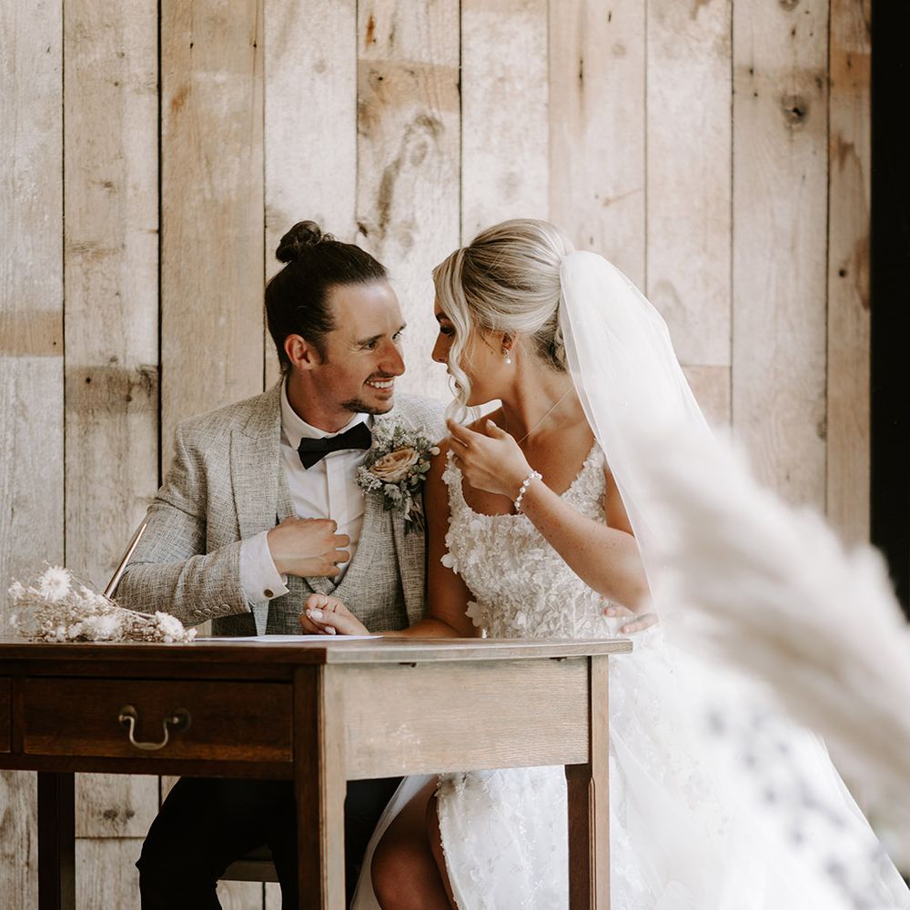 The bride and groom sit closely together and speak as they sign the wedding register together 