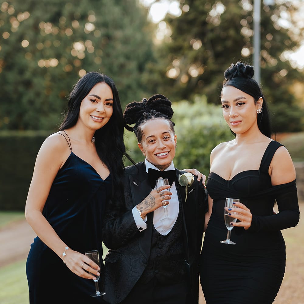 Bride in black snakeskin print tuxedo with black bowtie and white rose boutonniere standing with bridesmaids in mismatched black bridesmaid dresses 